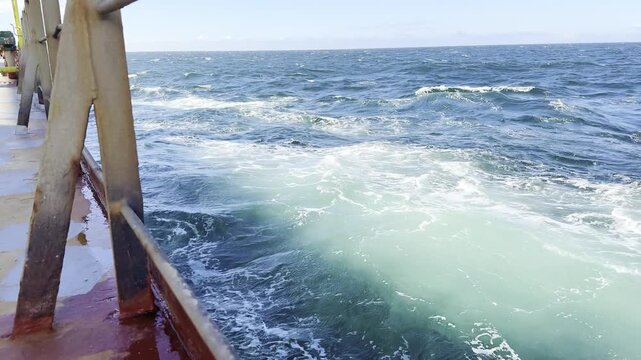 Cargo ship cutting through the ocean, creating white foam, sailing away. moderate ocean surface. view from the vessel deck.