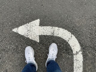 Fototapeta premium Person standing at a road junction with a white arrow pointing left, indicating a decision or a turn ahead.