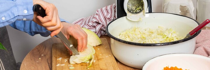 A 53-year-old man cuts cabbage with a knife while preparing to ferment cabbage and carrots. He stands in his kitchen near a vegetable grinder and a bowl of grated carrots, banner © serz72