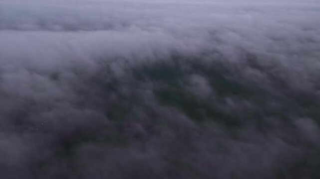 Airplane side window shot over a dense dramatic cloud layer revealing glimpses of green landscape and terrain below through gaps in the clouds at high altitude.