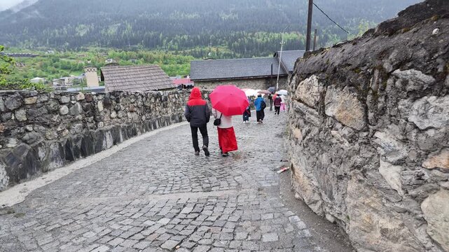 People walking on ancient wet stone cobblestone street with red umbrella in mountain village, historical architecture, travel tourism, Mestia, Svaneti, Georgia