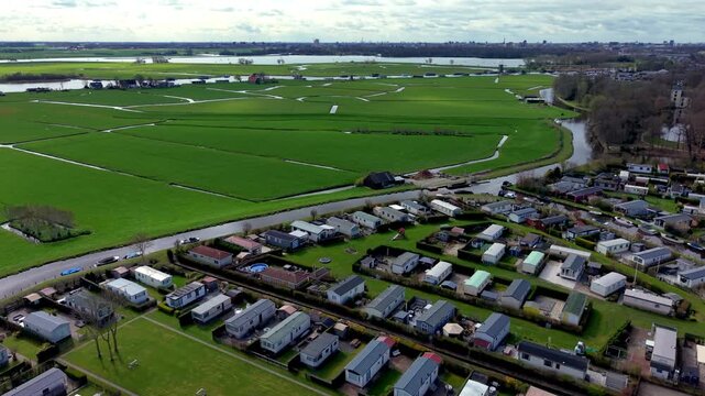 Aerial view of mobile home rows surrounded by green fields, intersecting water canals and distant urban skyline across flat rural landscape under partly cloudy sky