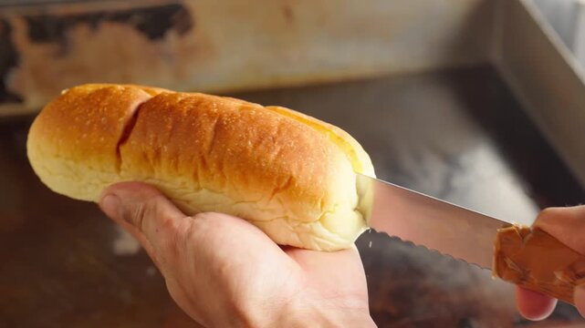 A chef using a bread knife to cut sandwich or hotdog bun, with hot flat top griddle on the background