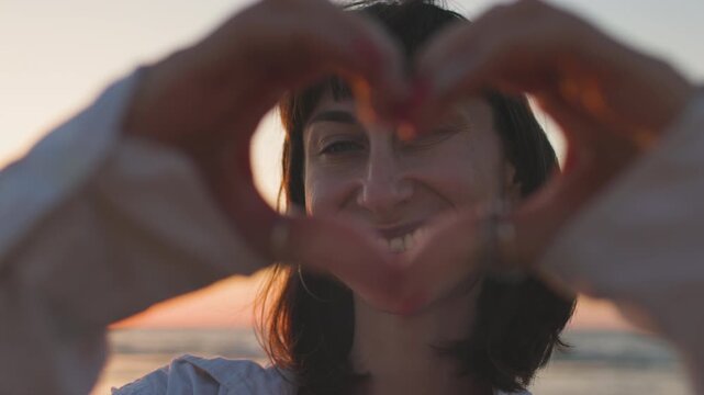 A close-up of a smiling woman on the beach, making a heart shape with her hands. A beautiful, cheerful woman laughs at the camera on the street. Concept of a healthy lifestyle, selflove.