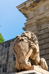 Obraz premium Close up view of small lion statue outside Mdina Gate, Malta in summer at sunset with limestone building behind on pillar below with lush green tree behind