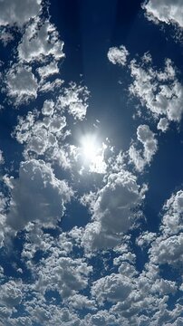 Bright sun shining through scattered fluffy clouds in a blue sky.