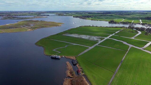 Aerial view of large waterbody bordered by green fields divided by narrow canals with small waterfront buildings, boats and flat rural landscape under partly cloudy sky