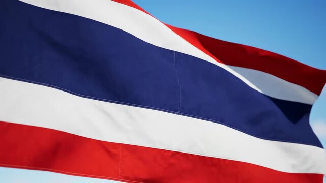 Thai flag with horizontal red, white and navy blue stripes waving in slow pan extreme close-up against bright blue sky with light clouds in daylight. thai national pride and royal monarchy identity