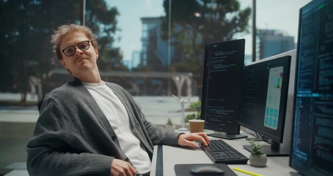 Young Software Developer Writes Application Code Across Multiple Displays in a Modern Tech Office, Then Turns to Smile at the Camera. Startup Developing New Mobile Apps. Working Using Computer