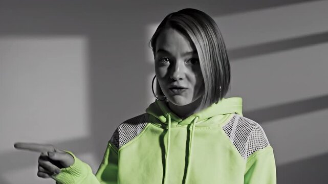 Young woman with short hair wearing a hoodie speaks and points to the side. Video sequence of casual communication and gesturing by a girl in interior setting.