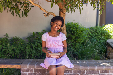 Naklejka premium African American child wearing pink dress sitting on brick bench under tree cradling bowl smiling