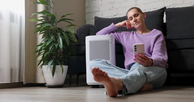 woman sitting on the floor by air purifier and using smart phone at home. air quality and microclimate