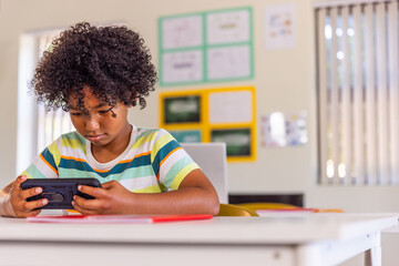 Naklejka premium African American boy child in classroom wearing striped shirt at desk holding device and red folder