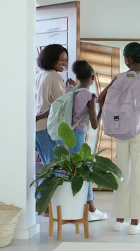 Vertical video: Holding door, mom sending girls for school in foyer, adjusting backpacks in mirror