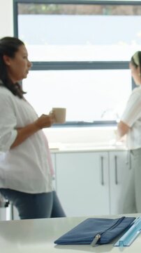 Vertical video: Friend entering, pregnant woman in white top sipping mug at home counter with pouch