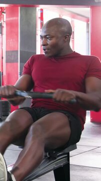 Vertical video: Rowing fit man in red shirt securing feet, doing strokes on rower at gym for cardio