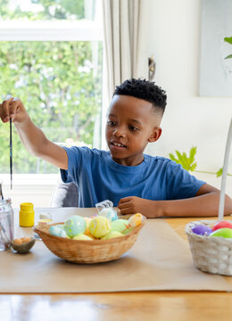 African American boy decorating eggs at table with wicker basket, yellow jar, brush, in blue tee