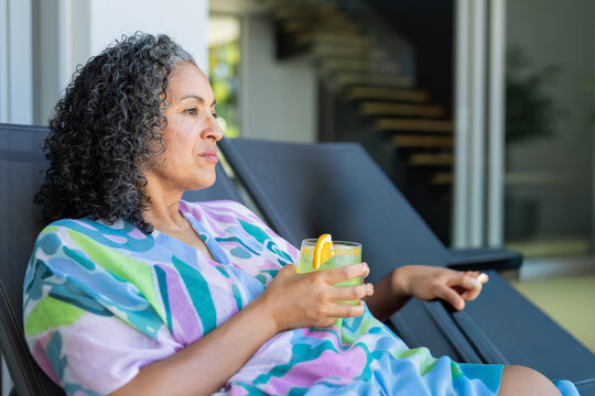 Middle-aged woman reclining on chair at terrace holding glass with pale green drink, copy space