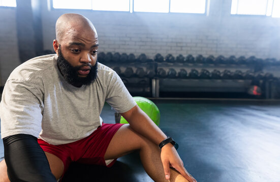 Adult African American man sitting on gym floor, wearing red shorts, resting beside medicine ball
