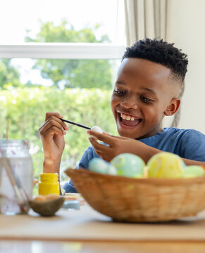 Youth African American boy painting egg with paintbrush at home table, woven basket nearby