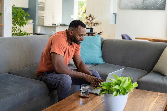 African American man sitting on sofa typing on silver laptop on coffee table in living room