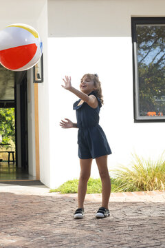 African American girl reaching, catching beach ball on brick patio at white house in navy romper