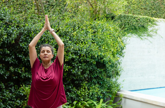 Mature woman standing poolside in garden, pressing palms above head, wearing maroon top and watch