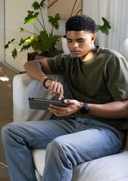 African American male sitting on light sofa tapping tablet in living room wearing olive tee