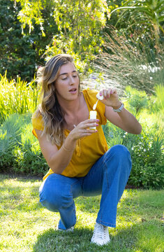 Woman squatting on lawn in garden wearing mustard top and jeans, holding bubble bottle and wand