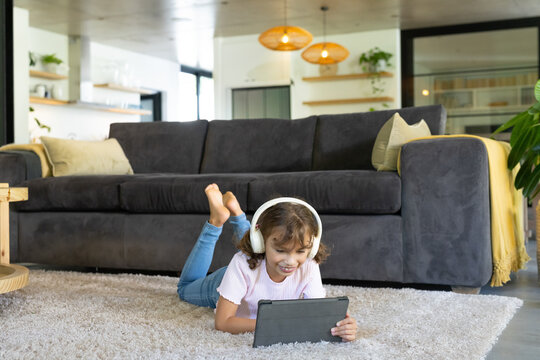African school-age girl lying on shag rug in living room wearing white headphones using tablet