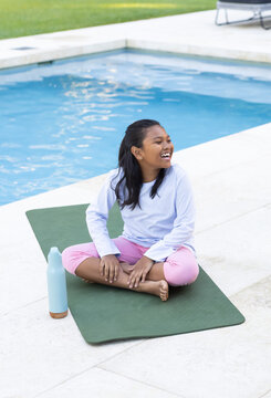 Female child sitting on green yoga mat beside pool, laughing near light blue bottle brown base