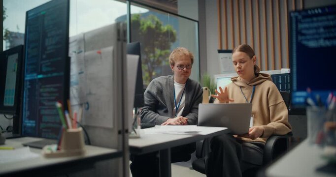 Two Software Engineers using Laptop Build in a Modern Tech Office, Coordinating Mobile App Ux, Ai Integration, Cybersecurity Checks. Two Young Professionals Talk While Working on Project Collaboration