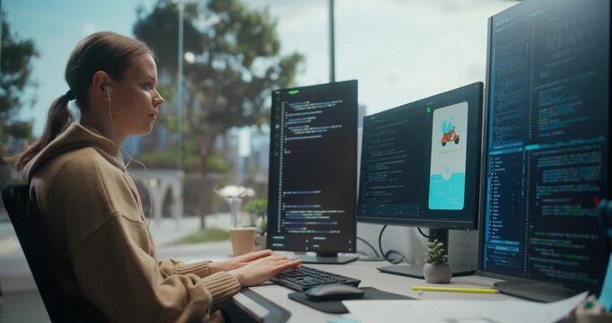 Young White Woman Software Engineer Wearing Earphones Typing Code on Multiple Computer Screens in Bright Modern Office, Developing Mobile Delivery Application Interface and Programming System