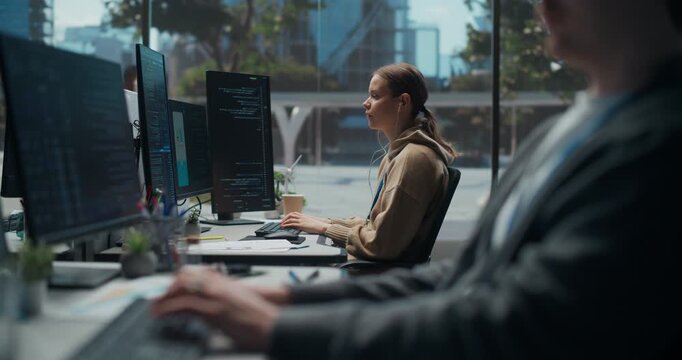 Focused Young White Female Programmer in Earphones Types Code on Dual Displays, Developing a Secure Mobile App in  Office While a Male Colleague Works Nearby, Tech Teamwork.
