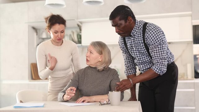 Thoughtful old woman compiling testament while middle-aged man and woman competing with each other in the kitchen