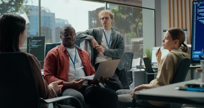 Black Male Software Engineer Holds Laptop and Leads Team Meeting as Diverse Developer Colleagues Talk App Architecture, Coding, Cybersecurity Interface Design in Tech Office.