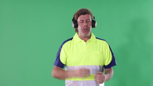  Adult male worker wearing reflective polo shirt stands against green screen, wearing headphones, moving rhythmically and dancing