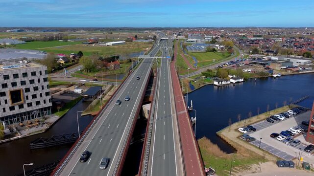 Aerial view of highway bridge crossing canal with parallel bike path, modern buildings, parking lot and mixed urban‑rural landscape extending across flat developed area
