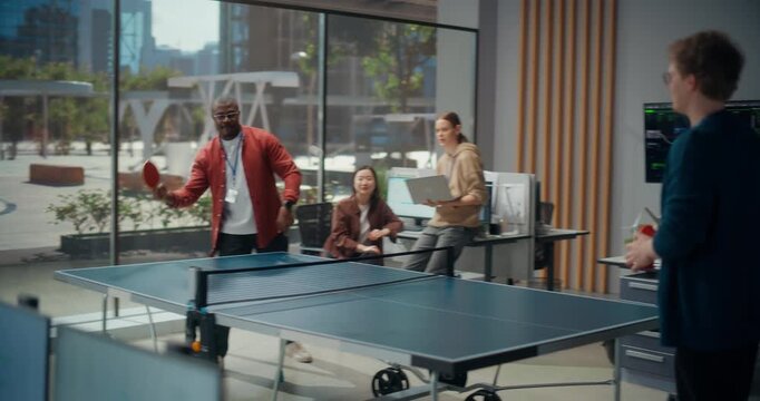 Tech Team Enjoys Friendly Table Tennis Match in Bright Startup Office. Black and White Male Developers Compete While Female Engineers Cheer, Holding Laptop Near Code-filled Monitors and Workstations.