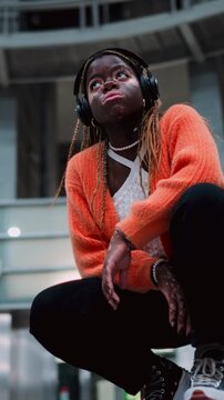 Stylish black female with vitiligo skin enjoying song beat in wireless headphones. Attractive African American listen to music from online playlist through earbuds while sitting in subway. Slow motion