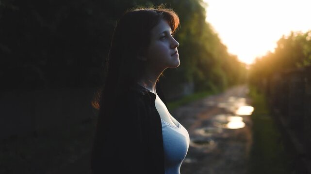 Beautiful woman stands outdoors looking back at the camera during a beautiful golden sunset. Dreamy backlighting, soft cinematic glow, peaceful atmosphere, warm sun rays, elegant portrait, summer