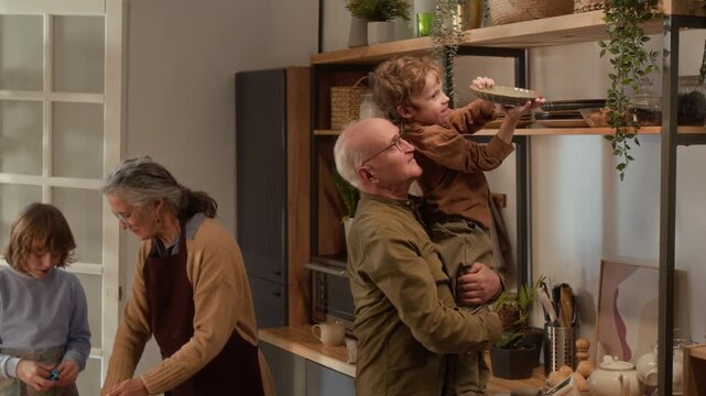 Tilt shot of senior Caucasian grandfather lifting young grandson to reach shelf while grandmother and older grandson bake. Ideal for promoting family values and multigenerational living