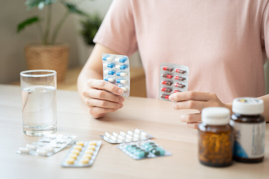 Close-up of a young Asian woman holding and comparing different medicine blister packs with a glass of water and pill bottles on the table