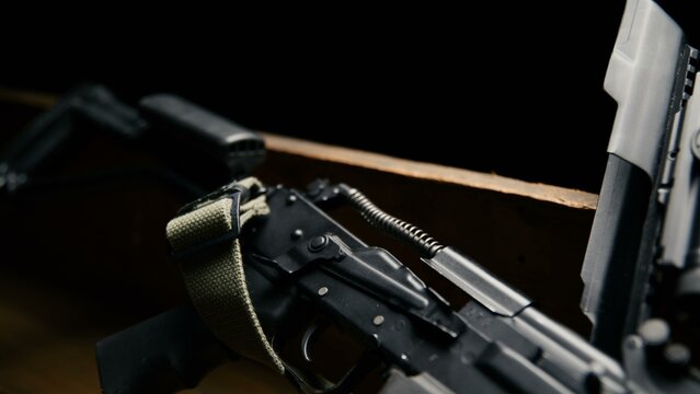 The matte black rifle receiver sits open with its internal recoil spring assembly exposed alongside an olive drab sling on a rustic wooden surface in this industrial hardware composition
