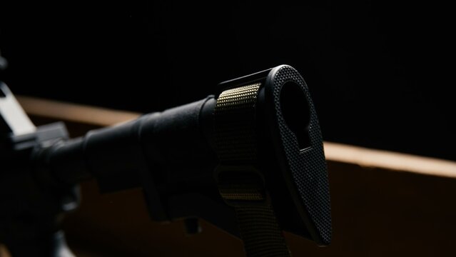 A close view of a black polymer rifle stock featuring a textured rear buttplate and an integrated tan nylon sling strap showcasing modular firearm furniture and support components
