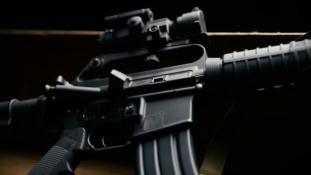 This side view displays a modular carbine rifle equipped with an optical sight and a durable sling placed on a wooden platform against a dark background, showcasing its tactical configuration