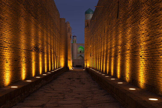 Illuminated brick walls of Ichan Kala in Khiva at twilight with minarets and tiled dome visible in the background. Perfect for travel guides, heritage publications, and tourism brochures.