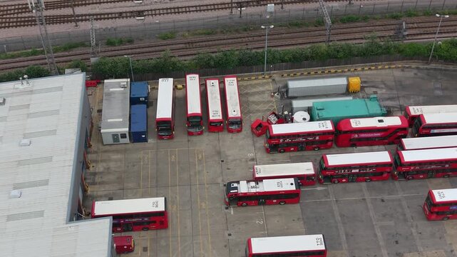 Lea Interchange bus garage seen from above as drone pans right over rows of red double-decker buses, depot yard, service bays and rail lines nearby in London, UK.
