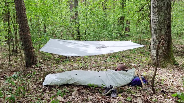 Tyvek bivy bag with a bearded man sleeping under a white poncho tarp in wild forest