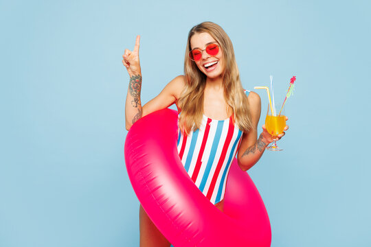 Young woman wear striped bikini swimsuit sunglasses rest near hotel pool hold pink rubber ring drink citrus cocktail show v-sign isolated on plain blue background. Summer vacation sea sun tan concept